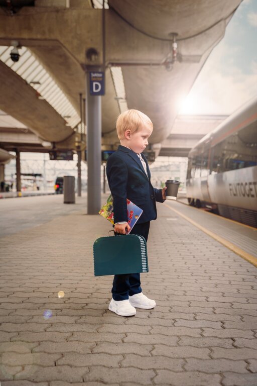 Barn i dress med kaffekopp, lekekoffert og magasin under armen. Venter på Flytoget
