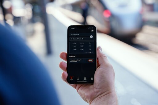 En iPhone i en hånd foran et flytog. På mobilen vises Flytogets app